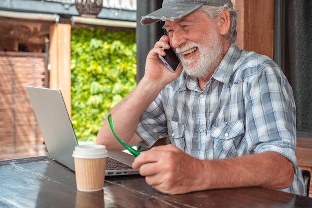 Photo cheerful senior man using laptop sitting at a wooden table while talking and laughing on mobile phone handsome elderly male browsing internet on laptop while drinking a coffee