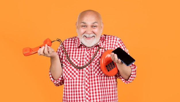 Веселый пожилой мужчина с ретро-телефоном и современным смартфоном на желтом фоне