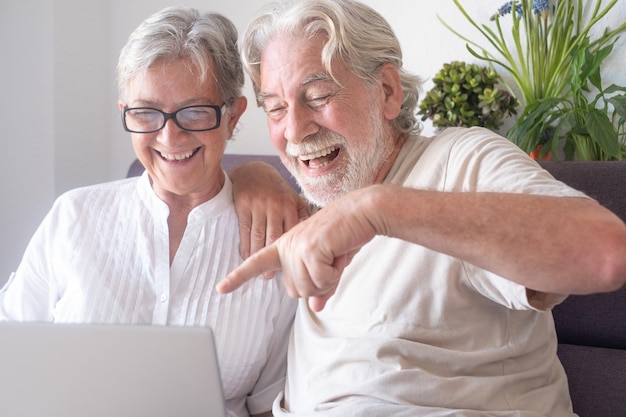 Cheerful senior white haired couple laughing in video chat with laptop Two caucasian retired person sitting on sofa at home enjoying tech and social