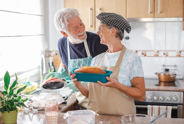 Cheerful senior woman holding her freshly baked homemade plumcake at her husband washing dishes