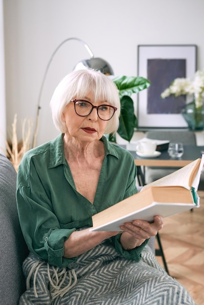 cheerful senior woman sitting on the couch reading a book at home