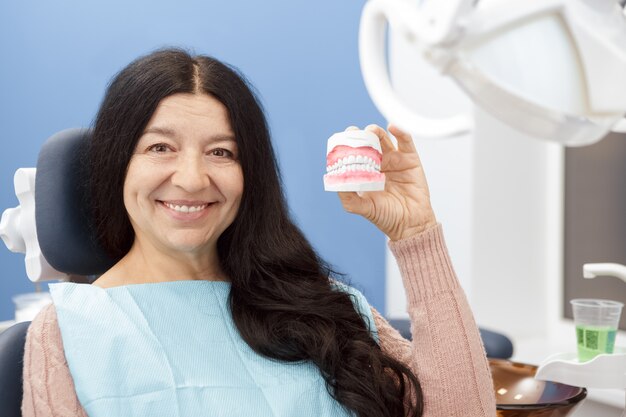 Cheerful senior woman smiling holding teeth mold at the dentist office