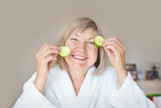 Cheerful senior woman with cucumber slices covering her eyes in front of her eyes in her bedroom. Spa beauty treatments at home, body care concept, organic cosmetics. Natural spa treatment.