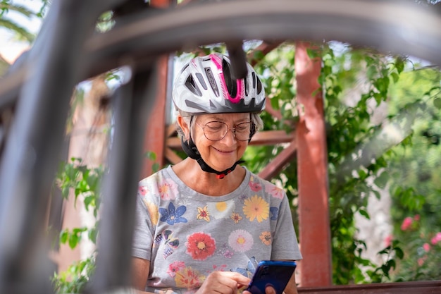 은퇴 후 건강한 생활 방식을 즐기는 노인 할머니가 휴대전화에 메시지를 작성하는 공공 공원에 앉아 헬멧과 자전거를 가진 쾌활한 노인 여성
