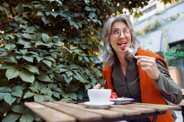 Cheerful senior woman with spectacles eats strawberry dessert sitting at table on outdoors cafe