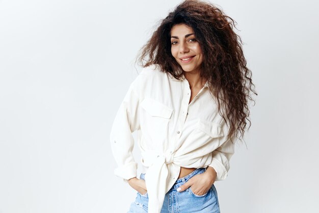 Cheerful sexy stylish curly pensive latin female lady with long\
afro hair in white shirt hold hands on jeans smiling at camera\
isolated at white studio background banner mockup copy space free\
place