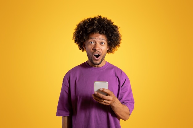 Cheerful shocked black adult curly man in purple tshirt with open mouth uses smartphone