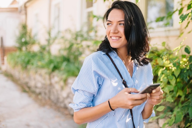 Ritratto allegro di donna attraente felice con il telefono cellulare guardando da un lato e in attesa di amici sulla strada della città giovane donna d'affari bruna che tiene smart phone copia spazio