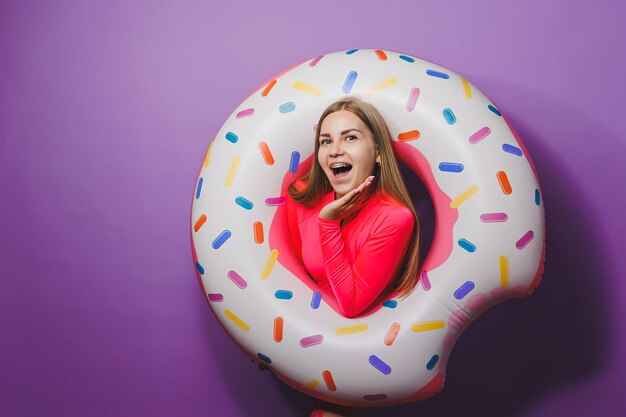 Cheerful slender woman with long hair holding an inflatable ring on a purple background Attractive girl in a pink swimsuit