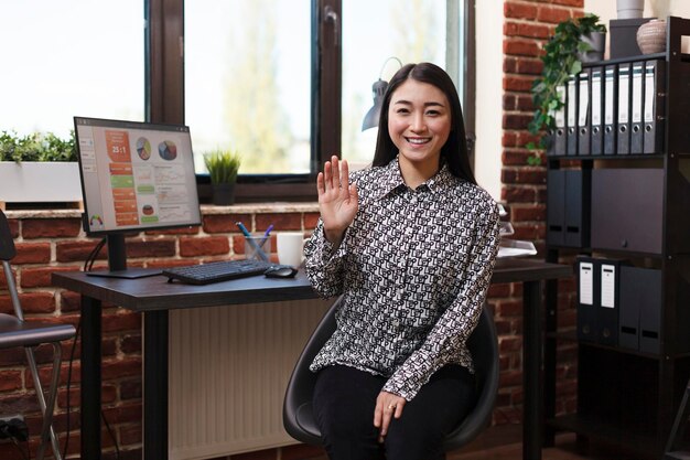 Cheerful smiling asian businesswoman sitting at desk in marketing agency workplace office while waving hand at camera. Happy financial company worker sitting in modern workspace interior.