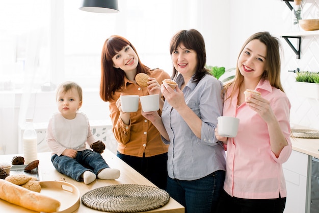 Giovani donne attraenti sorridenti allegre, donna invecchiata centrale e piccola neonata sveglia che mangiano i biscotti e i bigné e che bevono la cucina del caffè a casa. concetto di happy mothers day, cucinare insieme