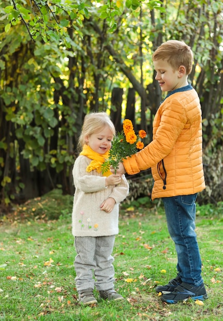 Cheerful smiling boy in yellow jacket presents flowers to little girl in yellow scarf in nature