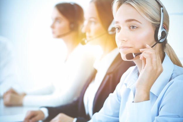Cheerful smiling business woman with headphones consulting clients. Group of diverse phone operators at work in sunny office.Call center and business people concept.