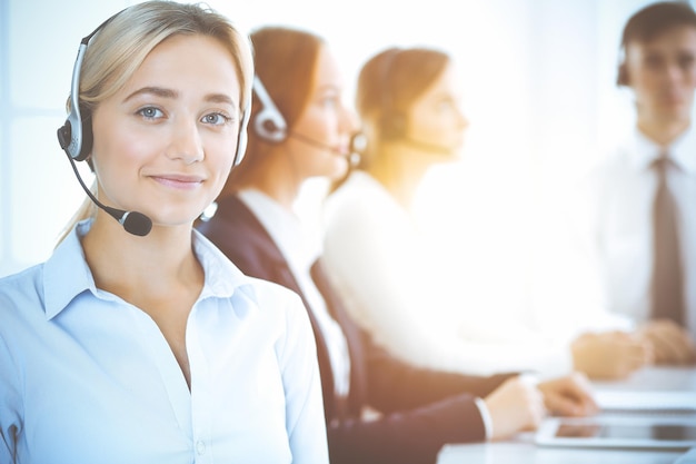 Cheerful smiling business woman with headphones consulting clients. Group of diverse phone operators at work in sunny office.Call center and business people concept.