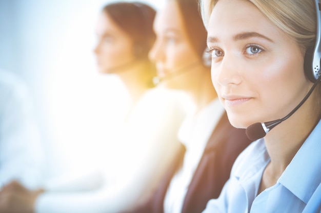 Cheerful smiling business woman with headphones consulting clients. Group of diverse phone operators at work in sunny office.Call center and business people concept.