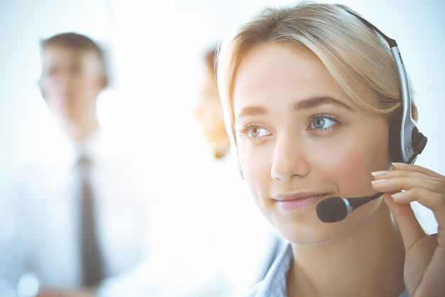 Cheerful smiling business woman with headphones consulting clients. Group of diverse phone operators at work in sunny office.Call center and business people concept.