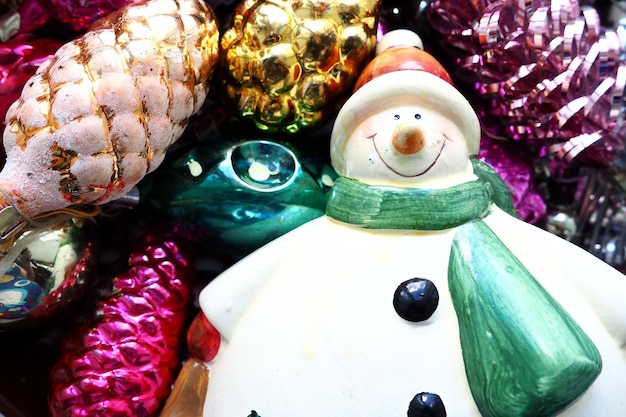 Cheerful smiling ceramic snowman in a green scarf and winter hat New Year and Christmas decorations tinsel Multicolored balls in the form of cones Festive interior decor New Year or Christmas