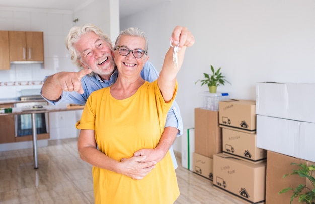 Cheerful smiling couple of senior people hugging holding the\
keys of the new empty apartment with moving boxes on the floor -\
concept of active elderly people and new beginning