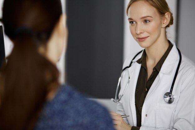 Cheerful smiling female doctor and patient woman discussing current health examination while sitting in clinic. Perfect medical service in hospital. Medicine concept.