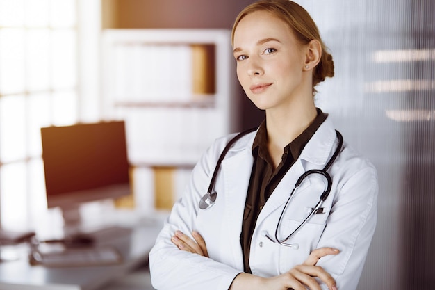 Cheerful smiling female doctor standing with arms crossed in clinic. Portrait of friendly physician woman. Medicine concept.