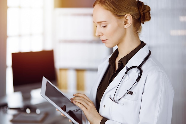 Cheerful smiling female doctor using clipboard in clinic. Portrait of friendly physician woman at work. Medical service in hospital. Medicine concept.