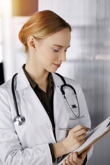 Cheerful smiling female doctor using clipboard in sunny clinic. Portrait of friendly physician woman at work. Perfect medical service in hospital. Medicine concept.