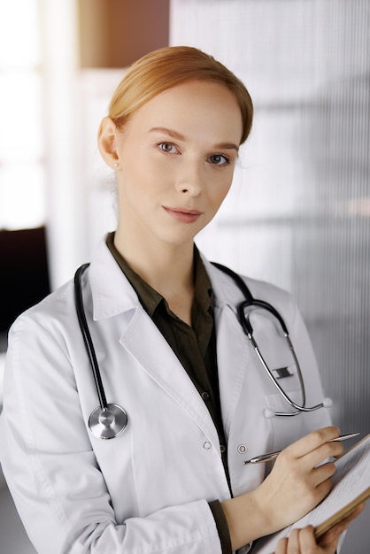 Cheerful smiling female doctor using clipboard in sunny clinic. Portrait of friendly physician woman at work. Perfect medical service in hospital. Medicine concept.