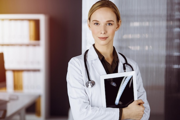 Cheerful smiling female doctor using tablet computer in sunny clinic. Portrait of friendly physician woman at work. Medicine concept.