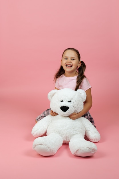 cheerful smiling girl in pink dress is sitting on floor near white teddy bear