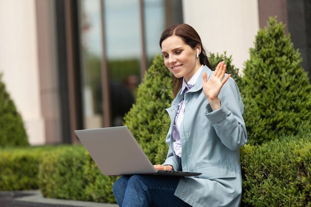 Cheerful smiling lady waves into the laptop screen in the city Video call conference lifestyle