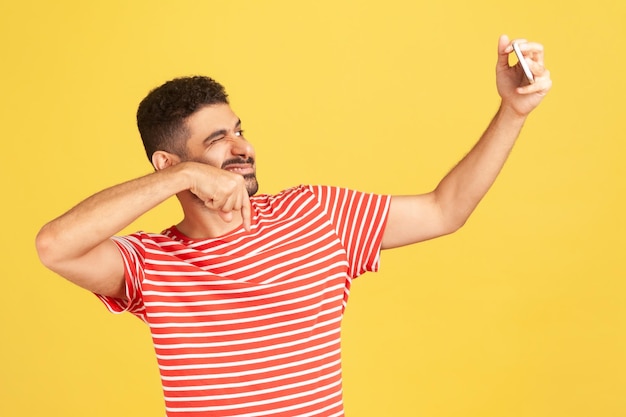 Cheerful smiling man blogger with beard in striped tshirt winking asking to subscribe looking at smartphone camera making selfie or recording video Indoor studio shot isolated on yellow background