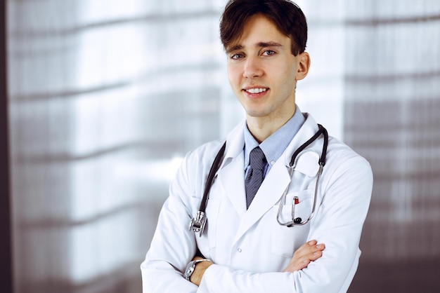 クリニックで腕を組んで立っている陽気な笑顔の男医師。病院で若い賢い医師との完璧な医療サービス。医学の概念。