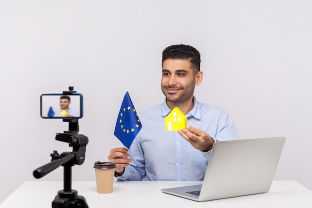 陽気な笑顔の男、不動産業者が紙の家と欧州連合の旗をカメラに見せ、ビデオを録画し、ヨーロッパでの購入または賃貸住宅についてのオンラインブログをストリーミングしています。スタジオショット分離