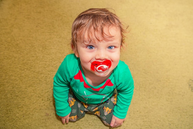 Photo cheerful smiling red-haired baby boy sits on the floor with a pacifier in his mouth