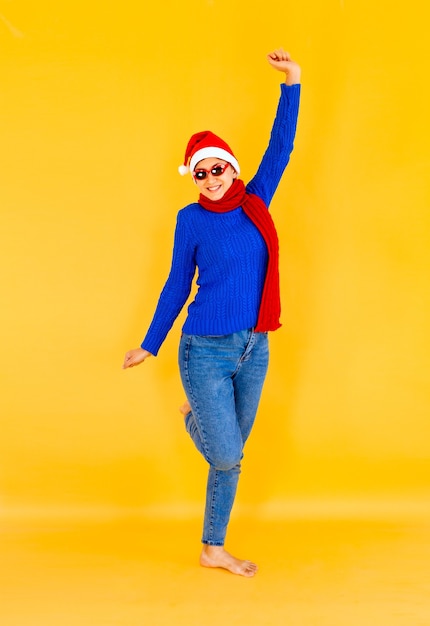 Cheerful smiling woman in blue sweater and santa hat raising her arm and leg, expressing happiness and success - studio shot isolated on yellow background. new Year