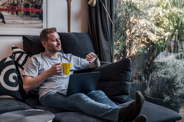 Cheerful smiling young adult man comfortably sitting at home on large sofa using laptop