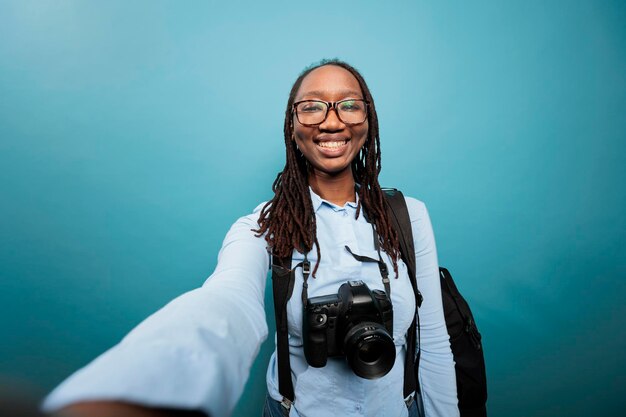 Cheerful smiling young adult photographer with DSLR camera taking selfie photo while standing on blue background. Creative joyful woman enjoying recording vlog video. Studio shot