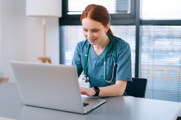 Cheerful smiling young female doctor in blue green medical uniform typing on laptop computer looking on screen sitting at desk near window in modern office of medic clinic.