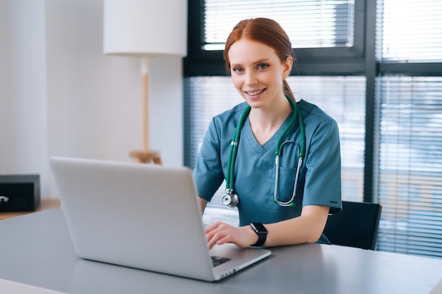 cheerful smiling young female doctor in blue green medical uniform typing on laptop computer