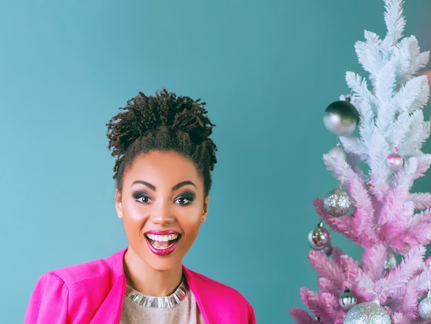 cheerful smiling young mixed rase woman in stylish fuchsia jacket near christmas tree