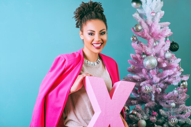 cheerful smiling young mixed rase woman in stylish fuchsia jacket near christmas tree