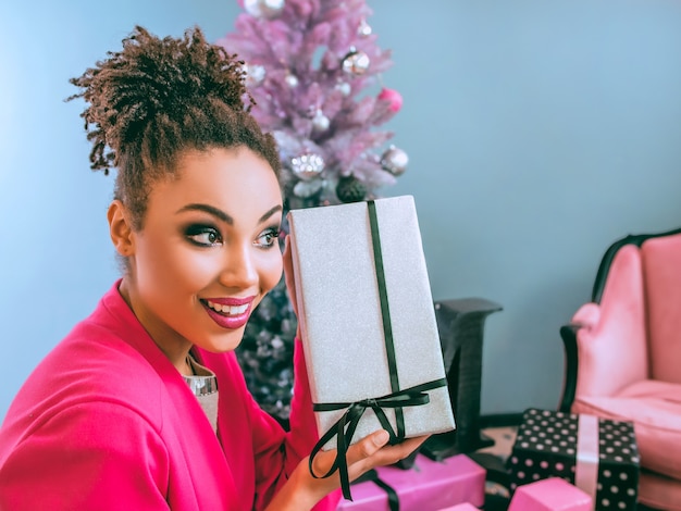 cheerful smiling young mixed rase woman in stylish fuchsia jacket near christmas tree