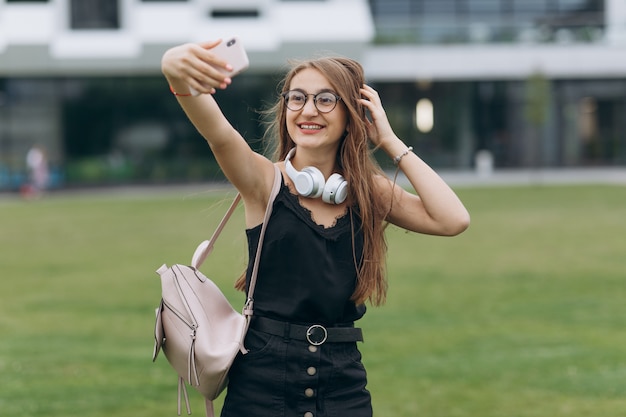 眼鏡をかけている長い髪の陽気な笑顔若い学生女子高生女性は、selfieを取ります。