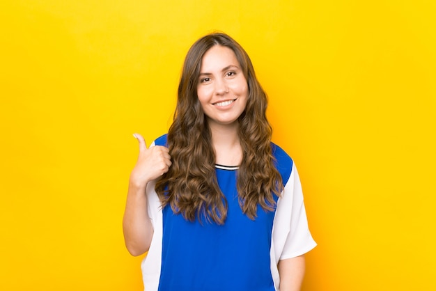 Веселая улыбающаяся молодая женщина в синей футболке показывает палец вверх на желтом фоне