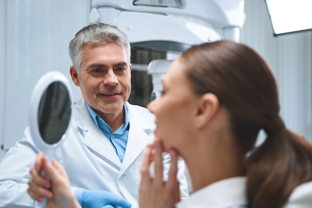 Cheerful specialist is looking at woman while she is looking into mirror after dental procedures