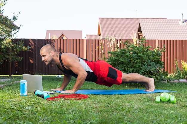 Cheerful sportsman  does pushups and watching online workout from laptop  in backyard