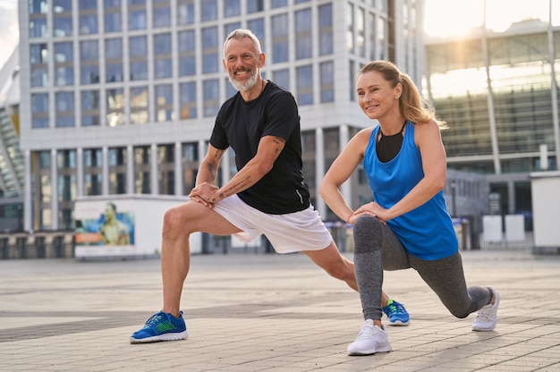 Cheerful sporty middle aged couple man and woman warming up getting ready for running together in