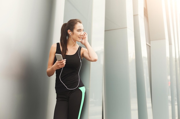 Cheerful sporty woman having rest after workout. Young fitness girl listening to music on smartphone, leaning on city construction, copy space