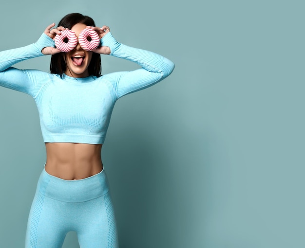 Cheerful sporty woman in sports leggings and top having fun closing her eyes with donuts