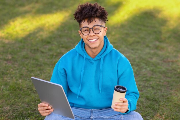 Cheerful student guy holds digital tablet and takeaway coffee outdoor